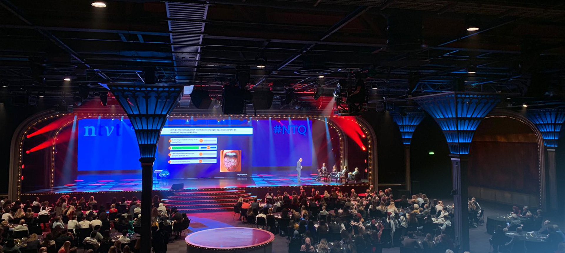 Volle zaal aan ronde tafels speelt een quiz mee, met de quizvraag op het grote scherm