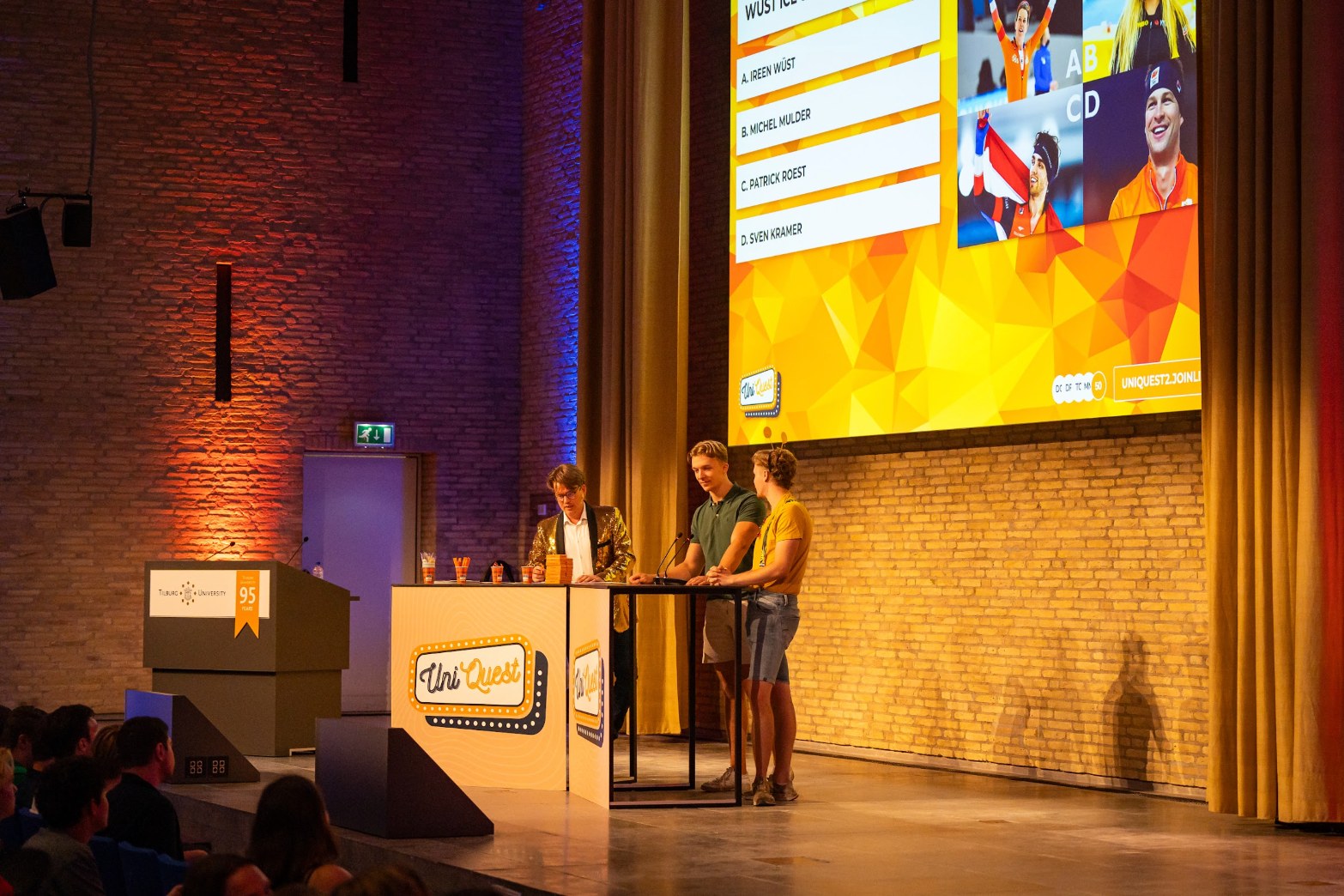 Jerre Maas en twee kandidaten aan de UniQuest-desk onder het scherm met een quizvraag